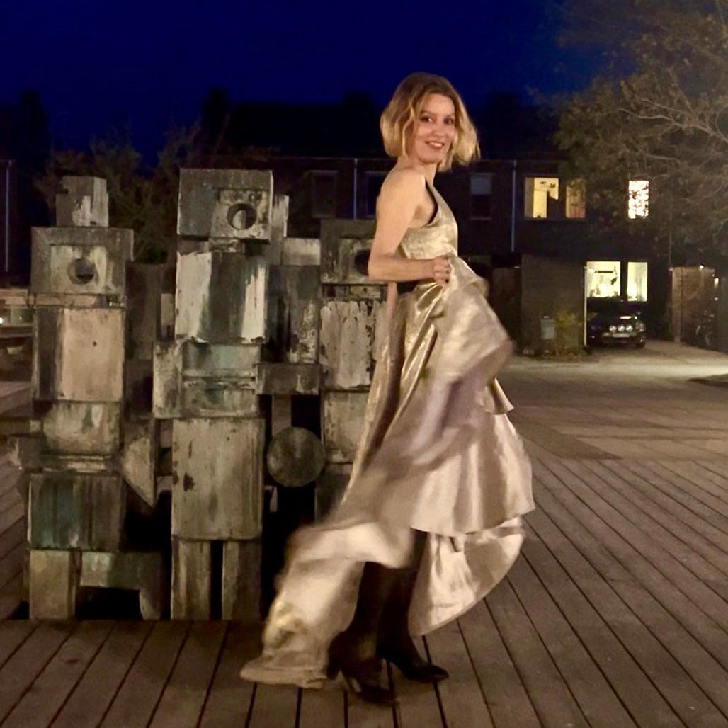A woman posing on a wooden boardwalk at night wearing a shimmering champagne-colored satin gown, standing next to a large weathered wooden sculpture.