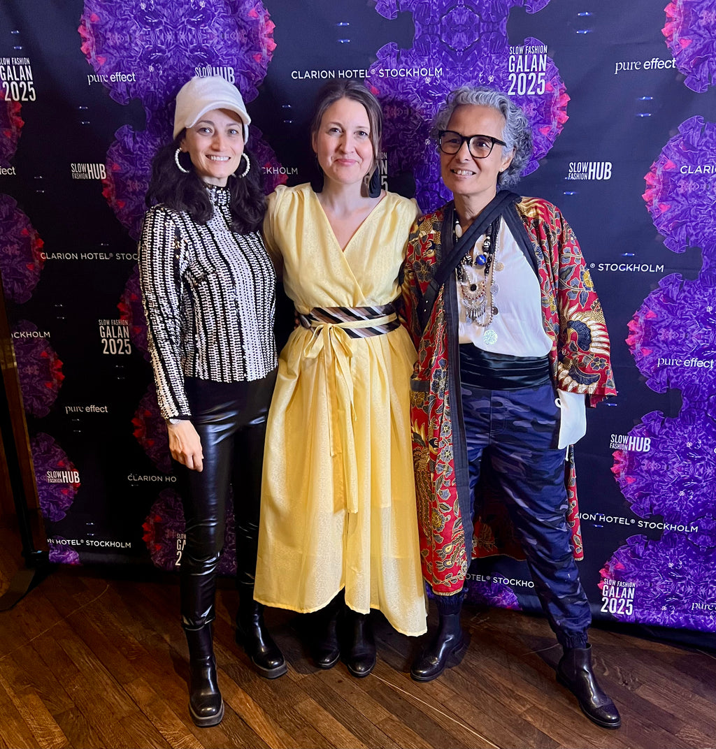 Three women posing together in front of a dark event backdrop printed with 'The Balan Stockholm 2025' and 'Clarion Hotel Stockholm' logos. The woman on the left wears a beige beanie, black-and-white striped long-sleeve top, black leather pants, and black boots.