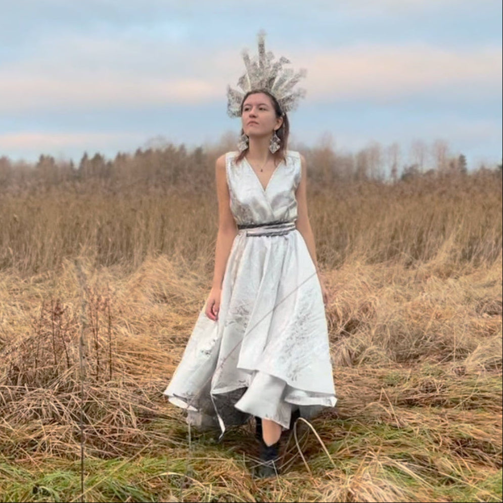 A woman stands in a grassy field wearing a sleeveless white dress with a silver waistband and a large feathered headpiece, photographed outdoors under a cloudy sky.