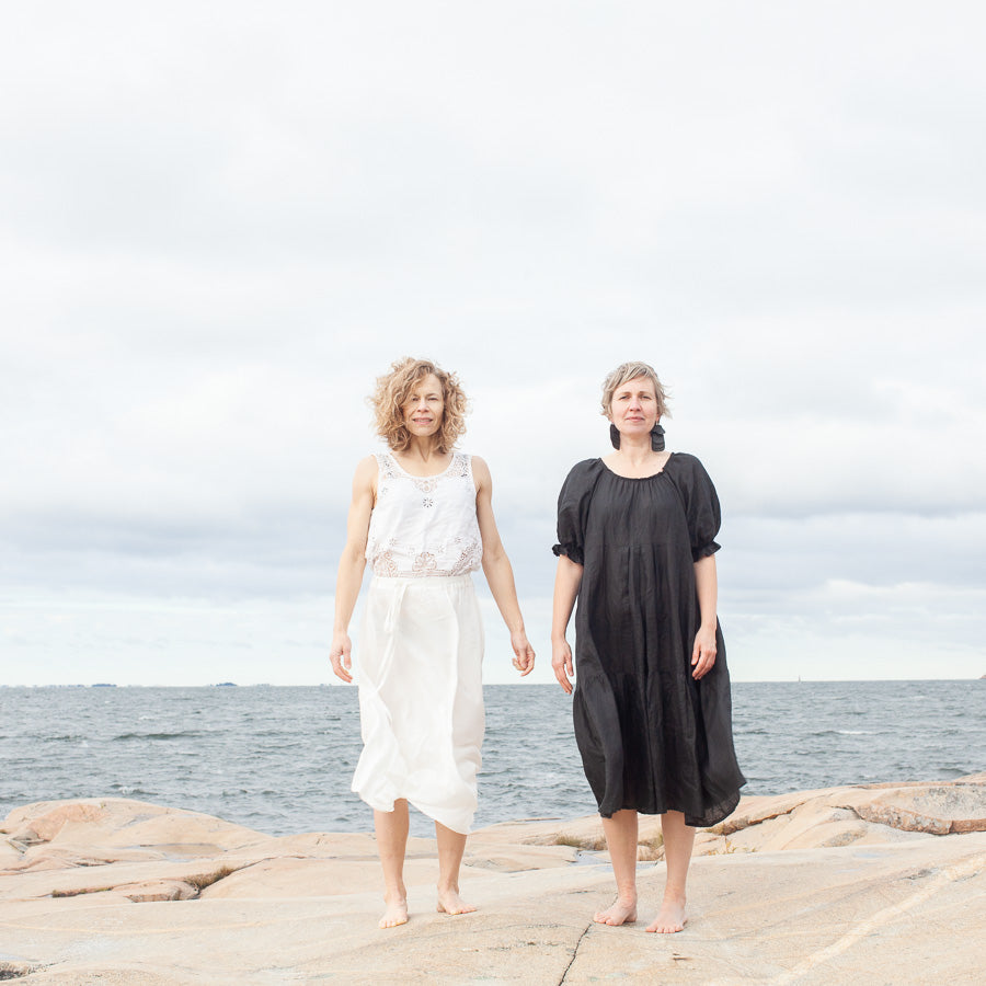 Two individuals standing on a rocky surface by the sea, wearing floor-length dresses. One dress is white and the other is black, with both individuals having curly hair.