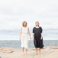 Two individuals standing on a rocky surface by the sea, wearing floor-length dresses. One dress is white and the other is black, with both individuals having curly hair.