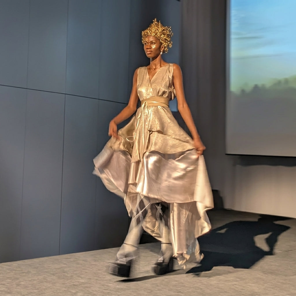 A model walks a runway wearing a shimmering metallic champagne dress with a high-low layered skirt, a matching waist belt, and a decorative gold headpiece in a fashion show setting.
