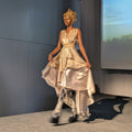 A model walks a runway wearing a shimmering metallic champagne dress with a high-low layered skirt, a matching waist belt, and a decorative gold headpiece in a fashion show setting.